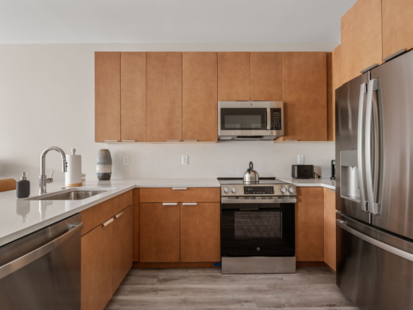 Modern kitchen with brown wood cabinets, stainless steel Refrigerator, range with oven, and over‑range microwave. White countertops and sink on left.