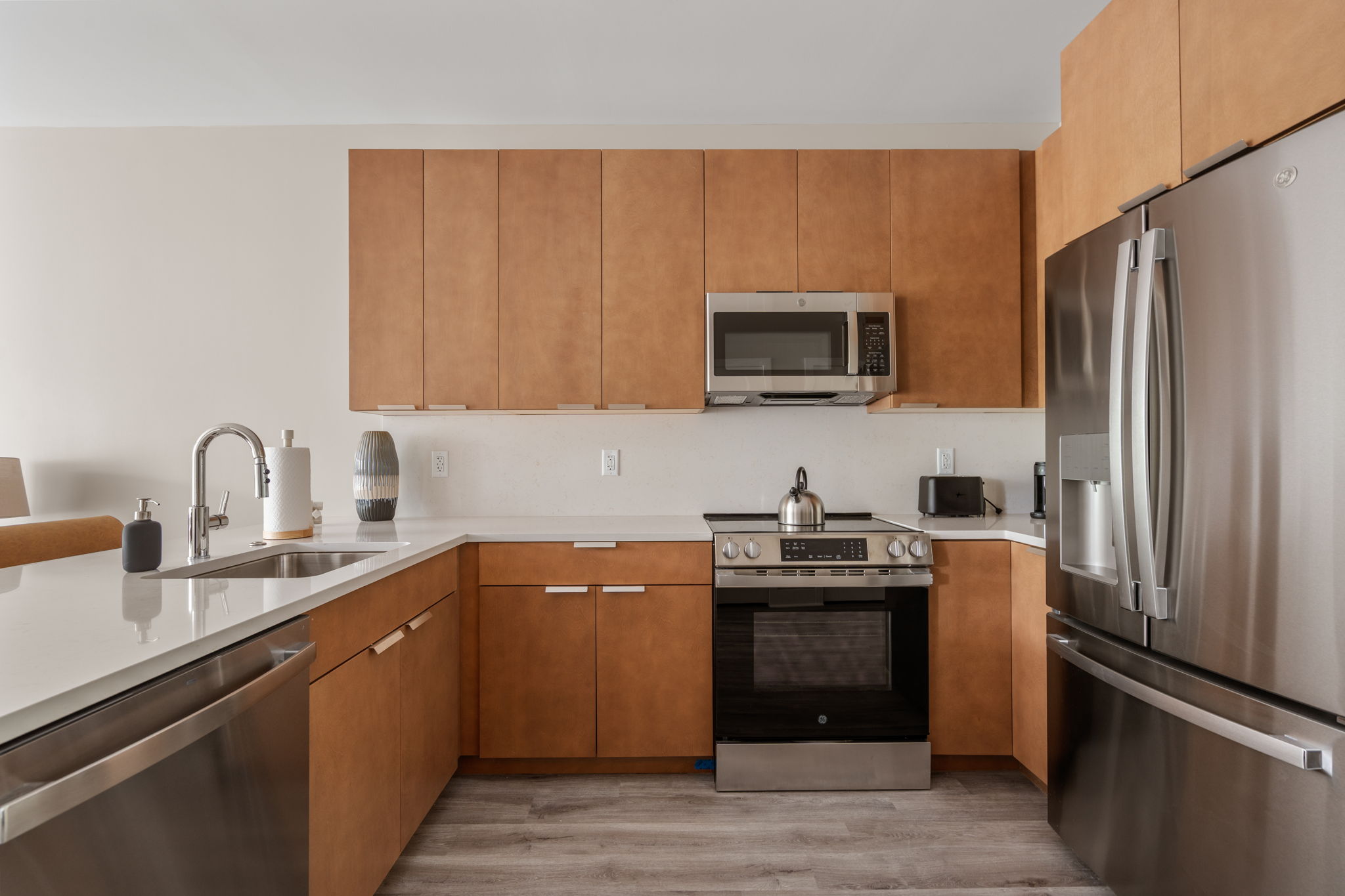 Dexter Street Commons, 1-Bed 2026-06-04 Modern kitchen with brown wood cabinets, stainless steel Refrigerator, range with oven, and over‑range microwave. White countertops and sink on left.