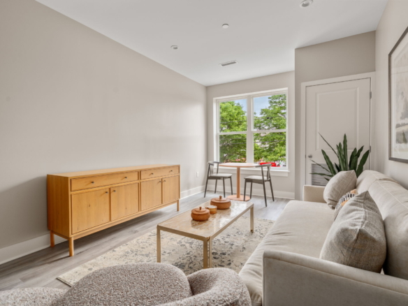 Bright living area with beige walls, a light wood sideboard, beige sofa, and a round table by a window overlooking green trees; plant in corner and decorative bowls on a coffee table.