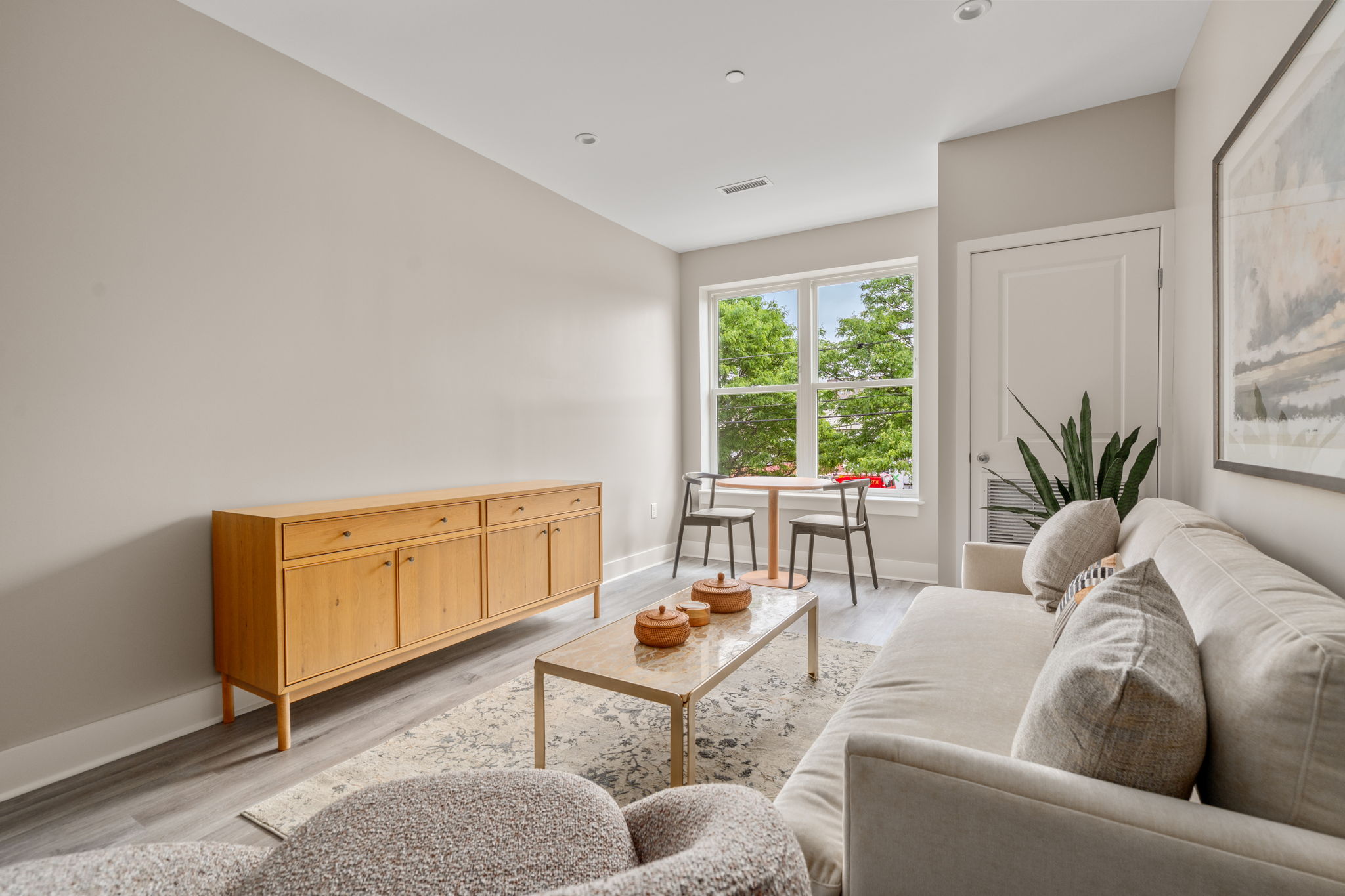 Dexter Street Commons, 1-Bed 2026-06-06 Bright living area with beige walls, a light wood sideboard, beige sofa, and a round table by a window overlooking green trees; plant in corner and decorative bowls on a coffee table.