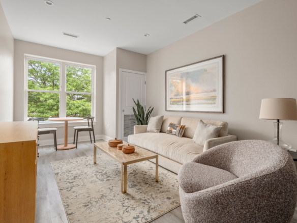 Bright living room with beige walls, a beige sofa, and a large framed landscape painting above the couch; sunlight from two windows to the left.