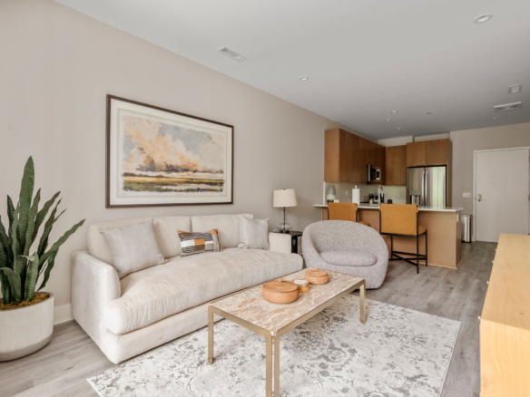 Open-concept living area with a light beige sofa, abstract wall art, and a tall potted plant; kitchen is visible in the background.