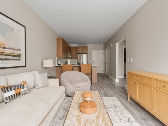 Open-concept living room with a beige sofa, patterned pillows, a marble coffee table, and a kitchen with wooden cabinets in the background.