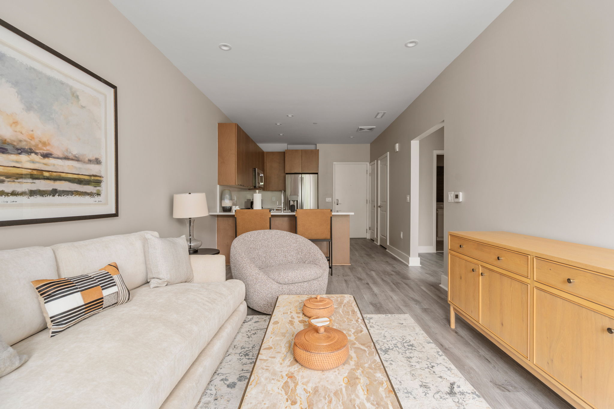 Dexter Street Commons, 1-Bed 2026-06-09 Open-concept living room with a beige sofa, patterned pillows, a marble coffee table, and a kitchen with wooden cabinets in the background.