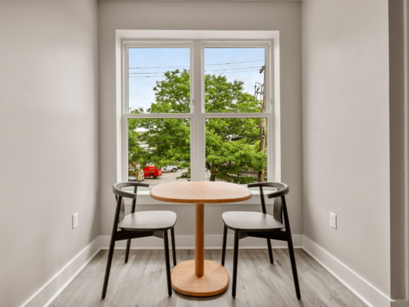 Narrow dining nook with a round wooden table and three dark chairs facing a large window with green trees outside.