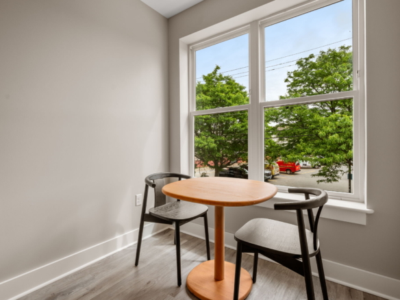 Corner breakfast nook with a round orange table and two black chairs by a large window; outside, green trees and parked cars are visible in daylight.