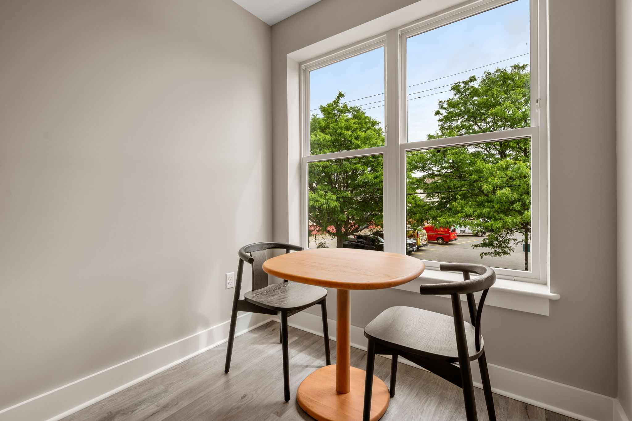 Dexter Street Commons, 1-Bed 2026-06-11 Corner breakfast nook with a round orange table and two black chairs by a large window; outside, green trees and parked cars are visible in daylight.