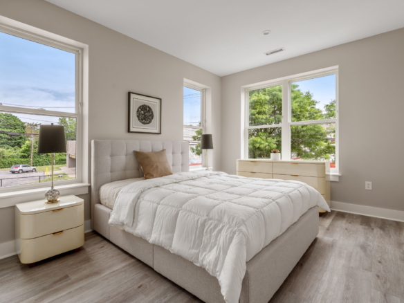 Bright modern bedroom with a tufted gray bed, cream nightstands, and large windows showing trees outside.
