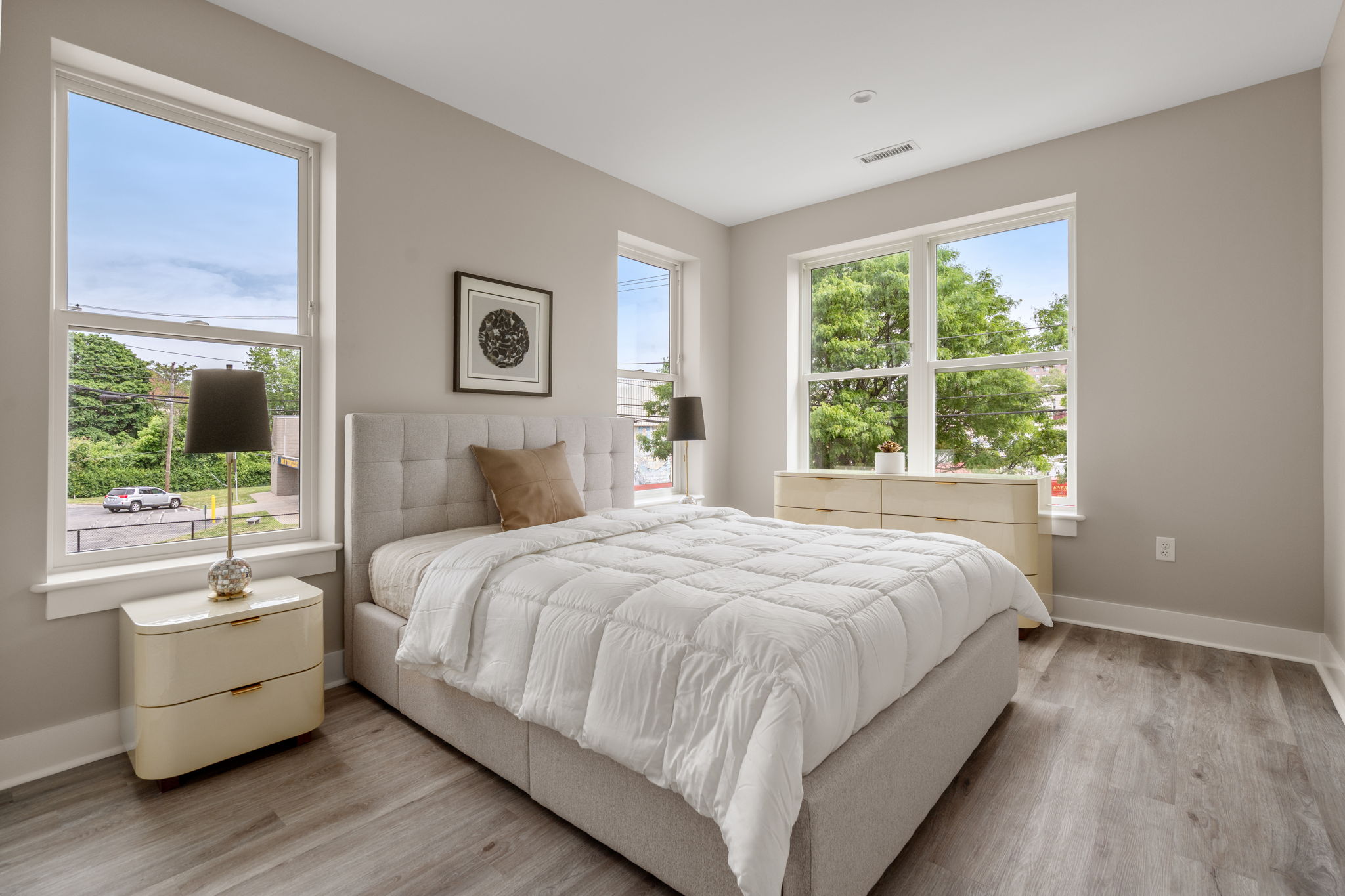 Dexter Street Commons, 1-Bed 2026-06-12 Bright modern bedroom with a tufted gray bed, cream nightstands, and large windows showing trees outside.