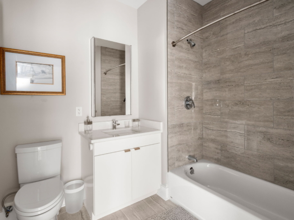 White modern bathroom with a white vanity, framed mirror, and a toilet on the left; a gray tiled bathtub/shower area on the right with a curved shower rod.