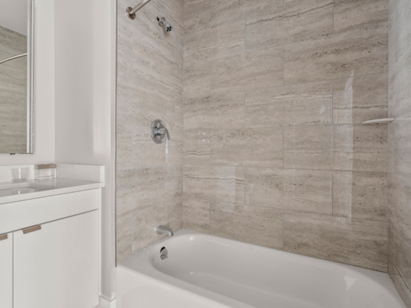 Modern bathroom with a white vanity and medicine cabinet next to a glass shower enclosure; beige-gray tiled walls surround a white bathtub edge.
