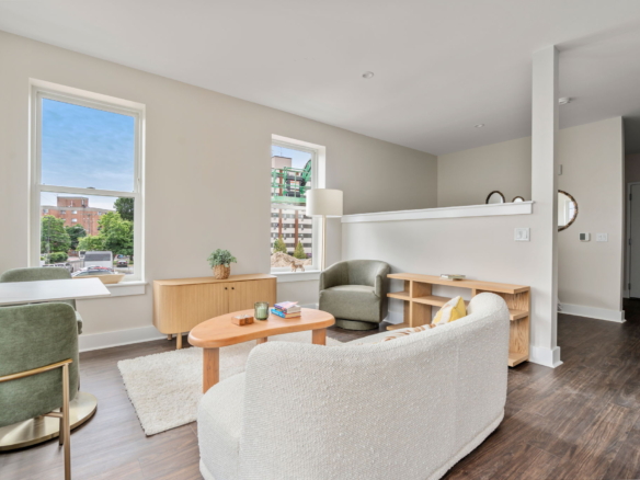 Bright, modern living room with large windows, light walls, and green accent chairs around wooden coffee tables.