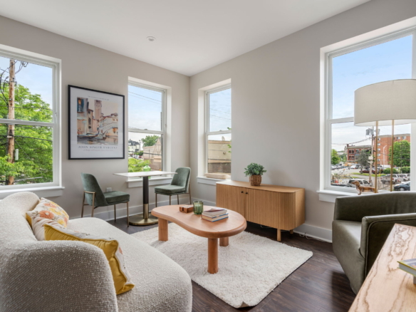 Bright living room with cream sofa, two green chairs, a round wooden coffee table, rug, and large windows showing trees and street outside.
