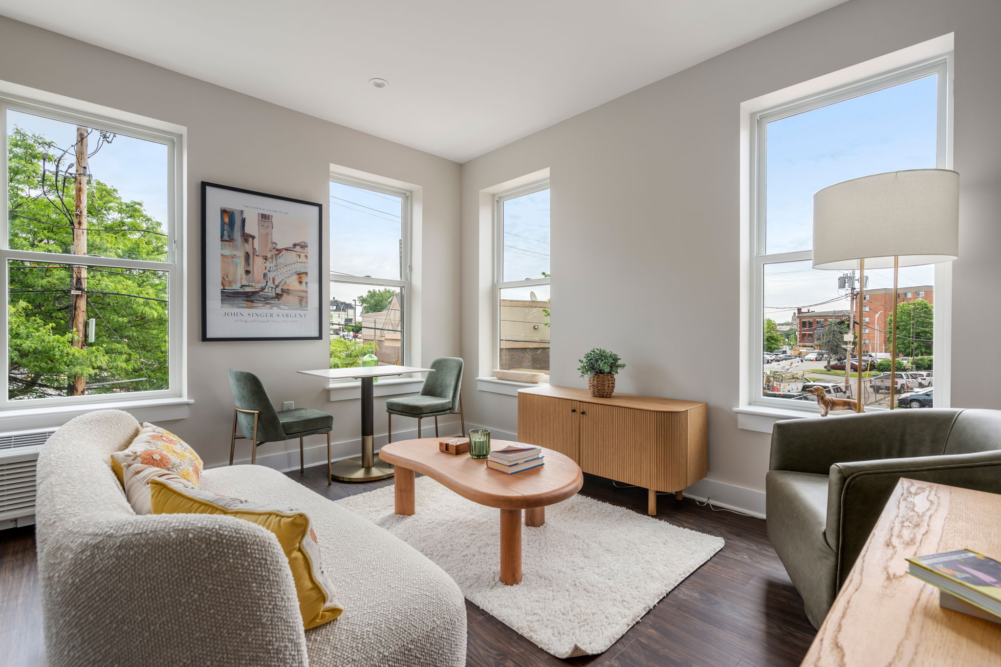 Bright living room with cream sofa, two green chairs, a round wooden coffee table, rug, and large windows showing trees and street outside.