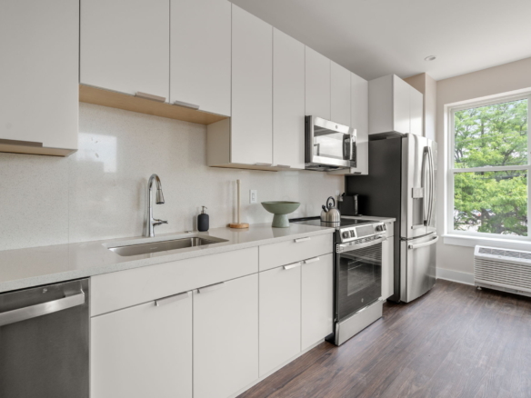 Modern white kitchen with stainless steel appliances and large window view of green trees outside, hardwood floors.