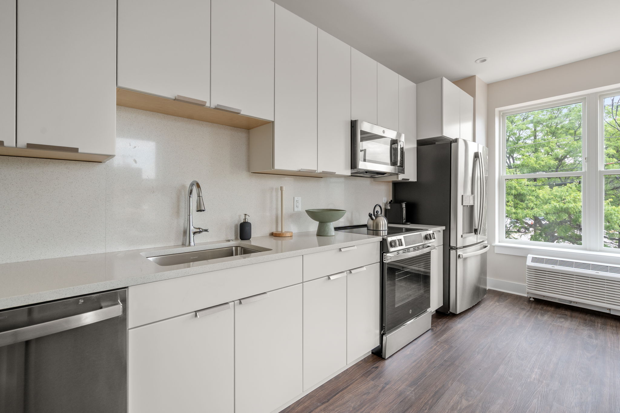 Modern white kitchen with stainless steel appliances and large window view of green trees outside, hardwood floors.