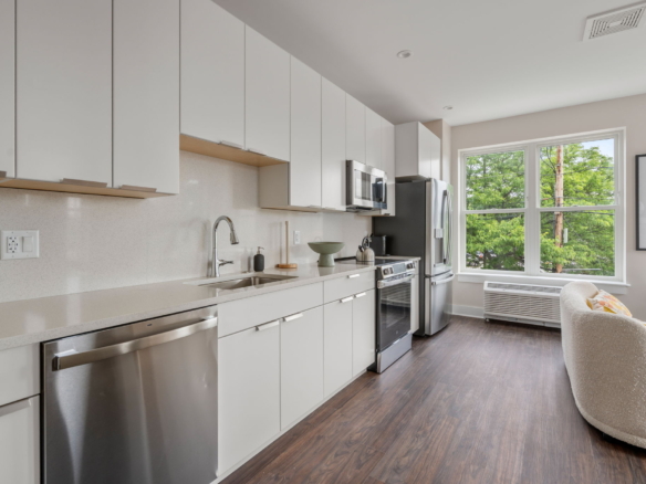 Bright, modern kitchen with white flat-panel cabinets, stainless steel appliances, and a large window letting in natural light over a dark wood floor.