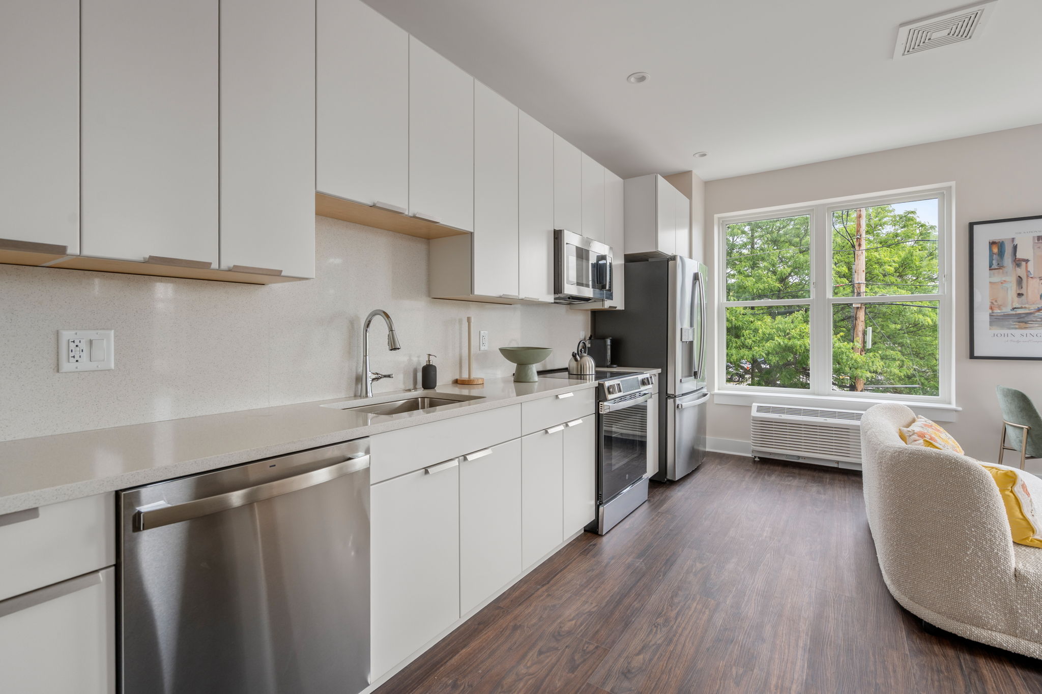 Bright, modern kitchen with white flat-panel cabinets, stainless steel appliances, and a large window letting in natural light over a dark wood floor.