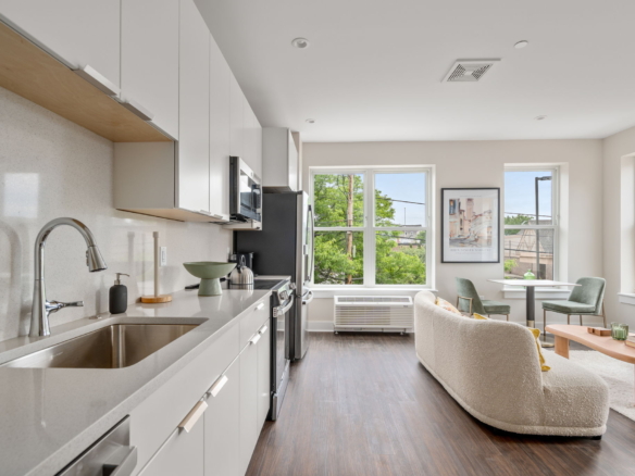 Bright open-plan kitchen with white cabinets and stainless-steel appliances, adjoining living area with large windows and a beige sofa.