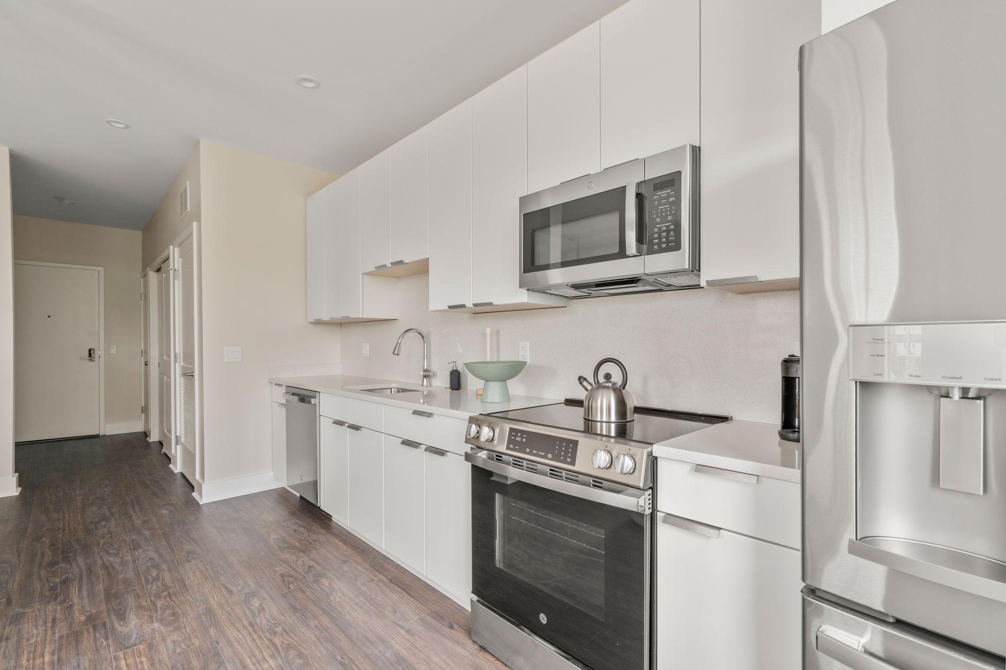 Modern white kitchen with stainless steel microwave and range, glossy cabinets, and a tall refrigerator along the right edge.
