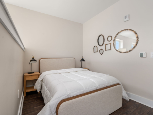 Bedroom with beige walls, upholstered bed, and two wooden nightstands with black table lamps; ornate mirrors on the right wall are arranged in varied shapes (oval, round, heart, rectangle).