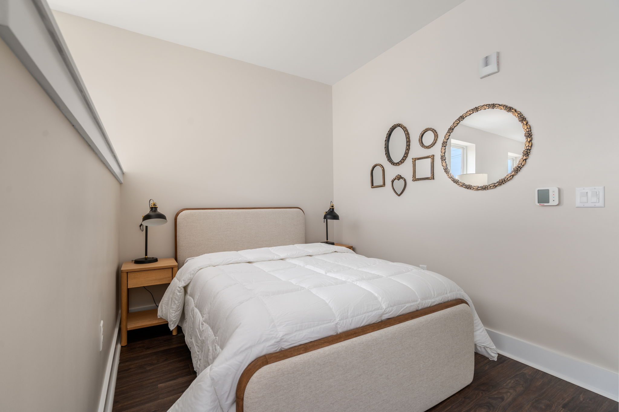 Bedroom with beige walls, upholstered bed, and two wooden nightstands with black table lamps; ornate mirrors on the right wall are arranged in varied shapes (oval, round, heart, rectangle).