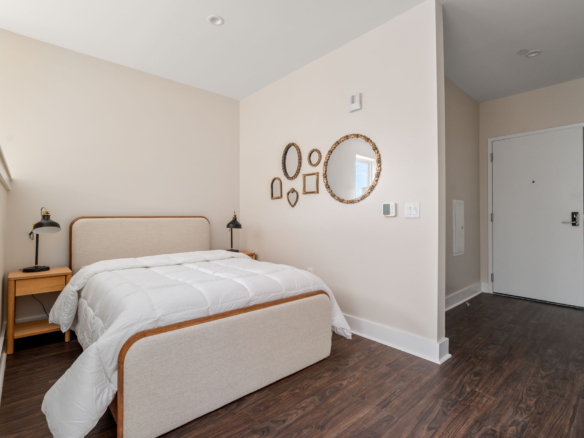 Neutral-toned hotel-style bedroom with a single beige headboard bed, white quilted bedding, and two wooden nightstands with black lamps. Wall decor includes nested oval mirrors. Doorway visible on the right.