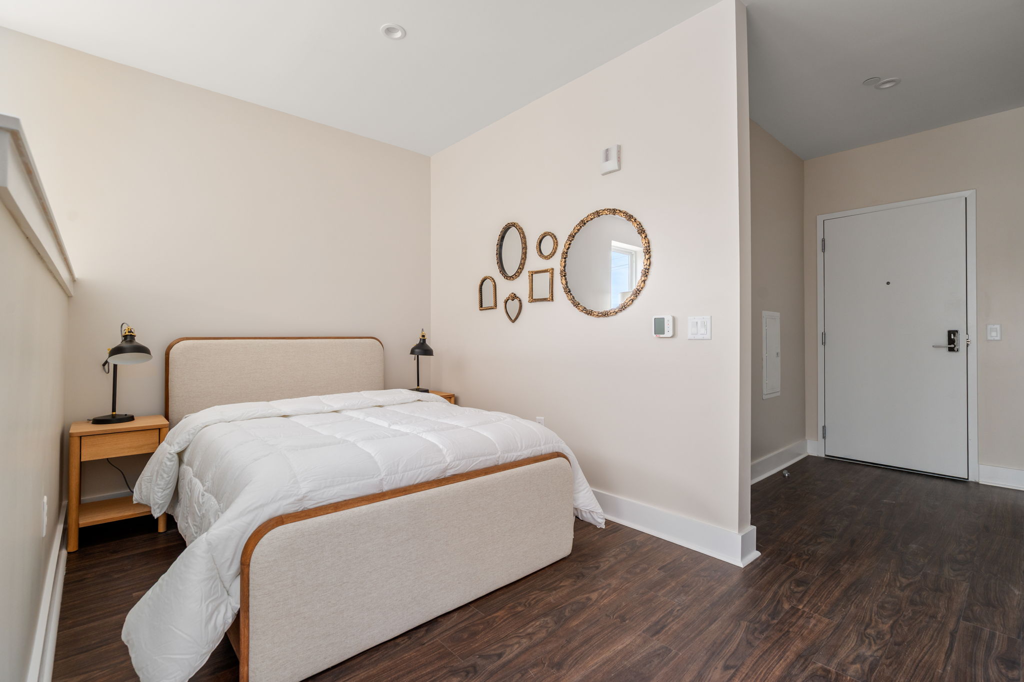 Neutral-toned hotel-style bedroom with a single beige headboard bed, white quilted bedding, and two wooden nightstands with black lamps. Wall decor includes nested oval mirrors. Doorway visible on the right.