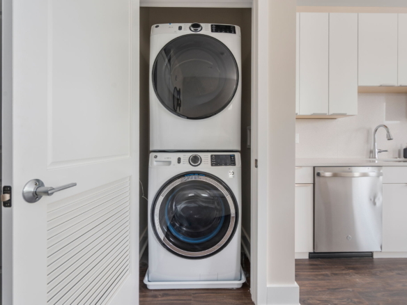 Stacked white front-loading washer and dryer in a laundry closet beside white kitchen cabinets.