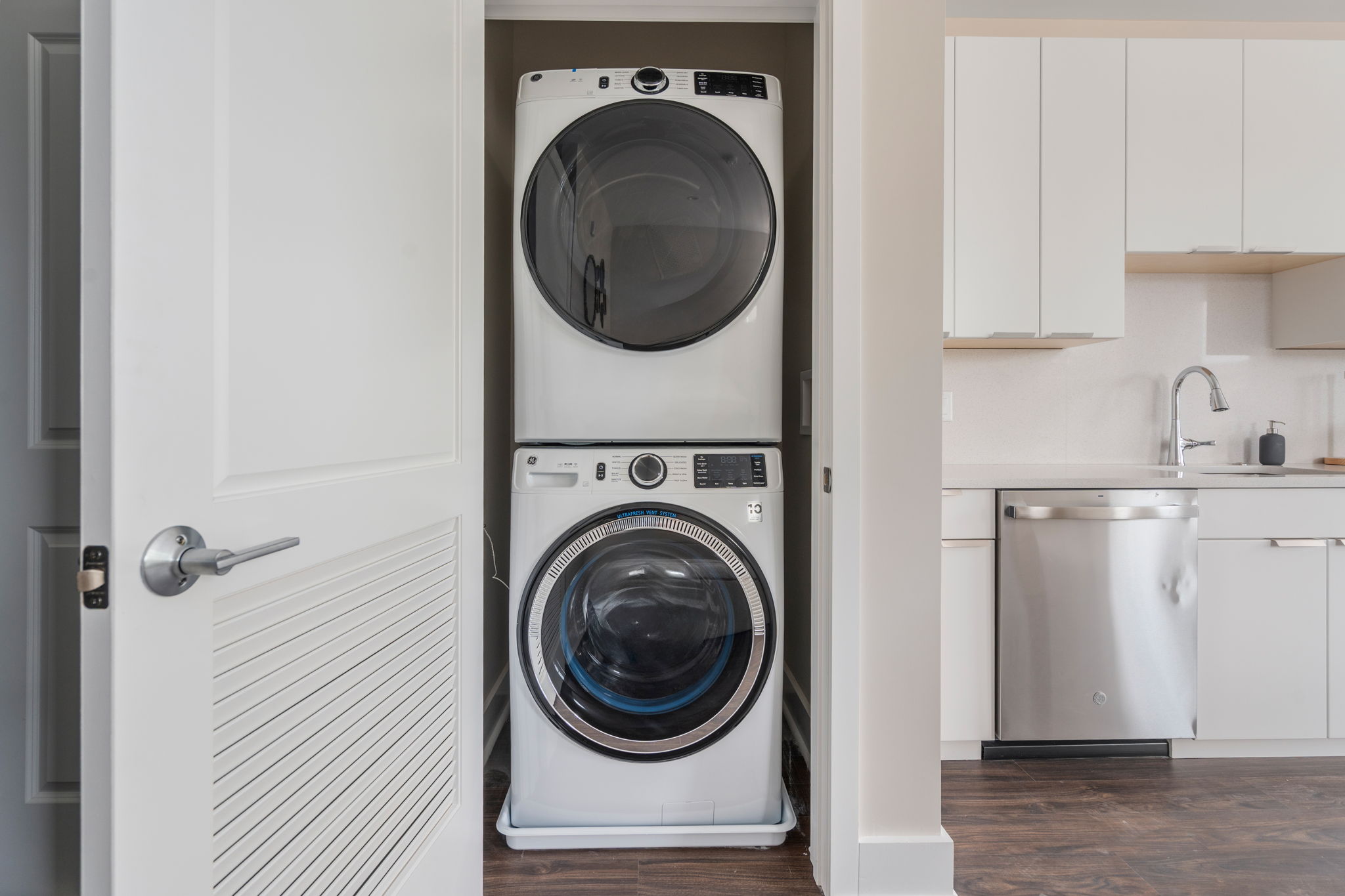Stacked white front-loading washer and dryer in a laundry closet beside white kitchen cabinets.