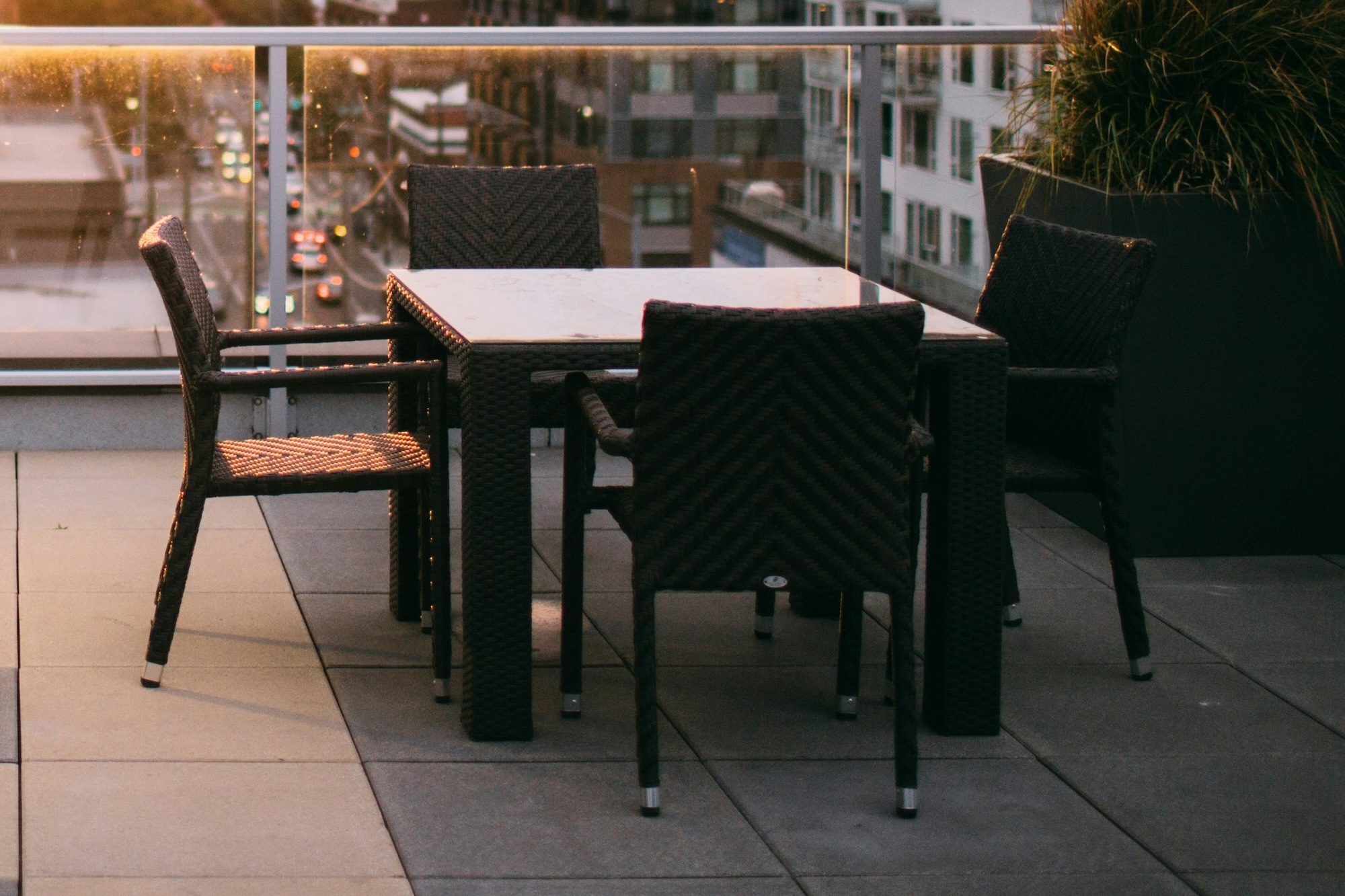 Wicker patio dining set on a rooftop terrace with a glass railing and city buildings in the background at dusk/purple hour.