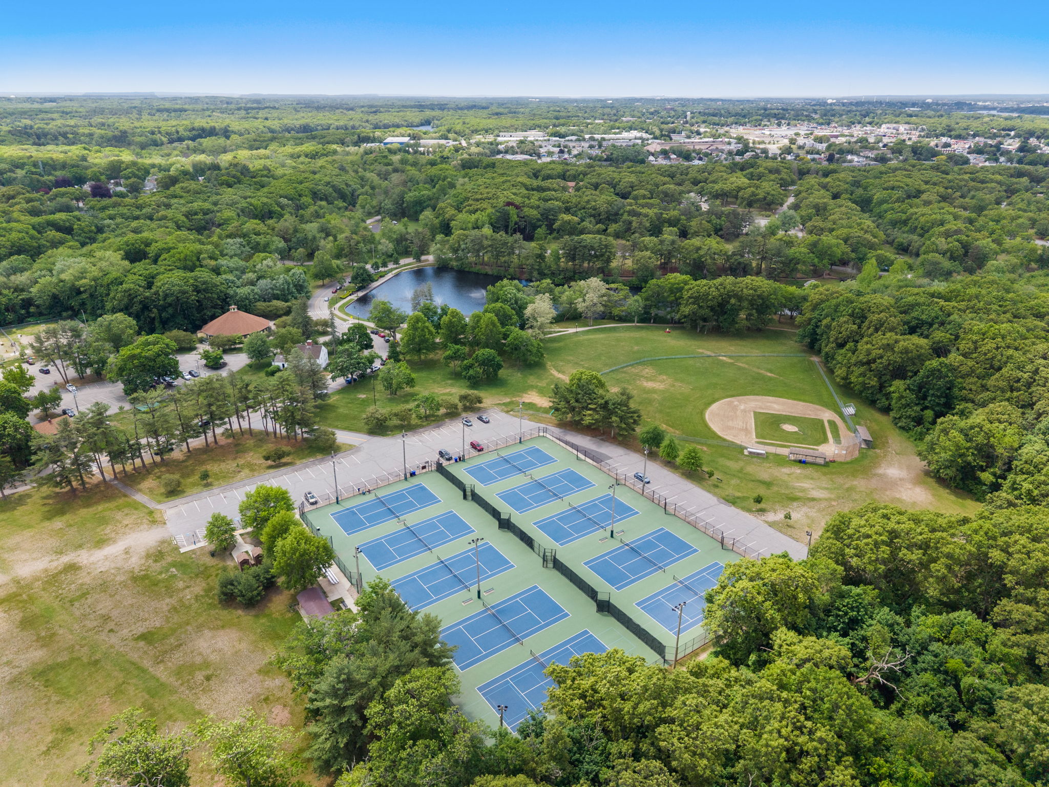 Aerial view of a large park with a pond, tennis courts in the foreground, and a baseball field nearby among green trees and open spaces