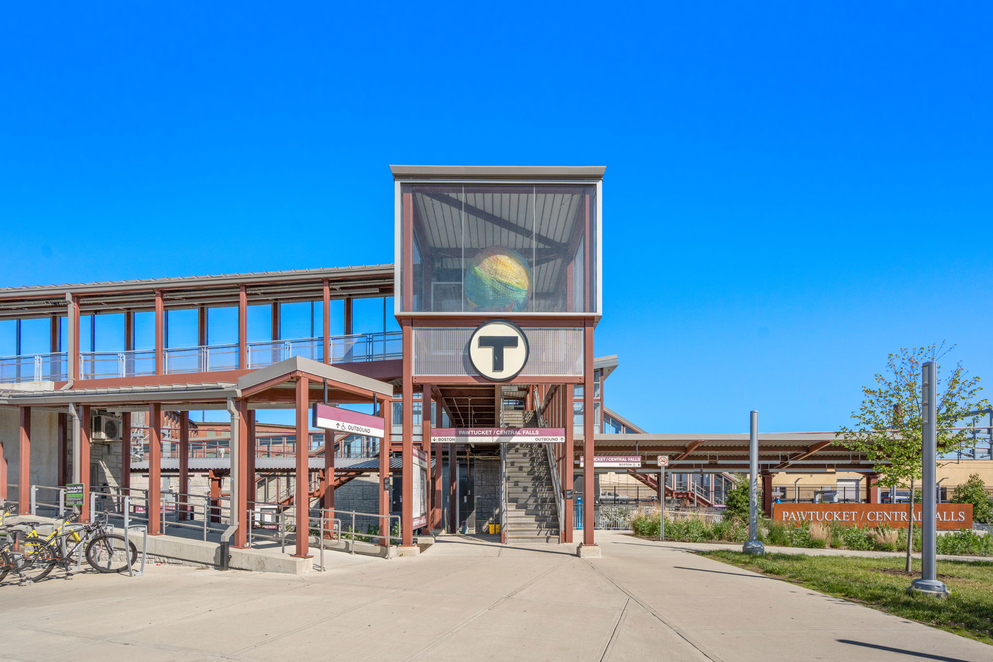 Transit station entrance with a glass-walled tower and a large T logo, bicycles on the left, under a clear blue sky.