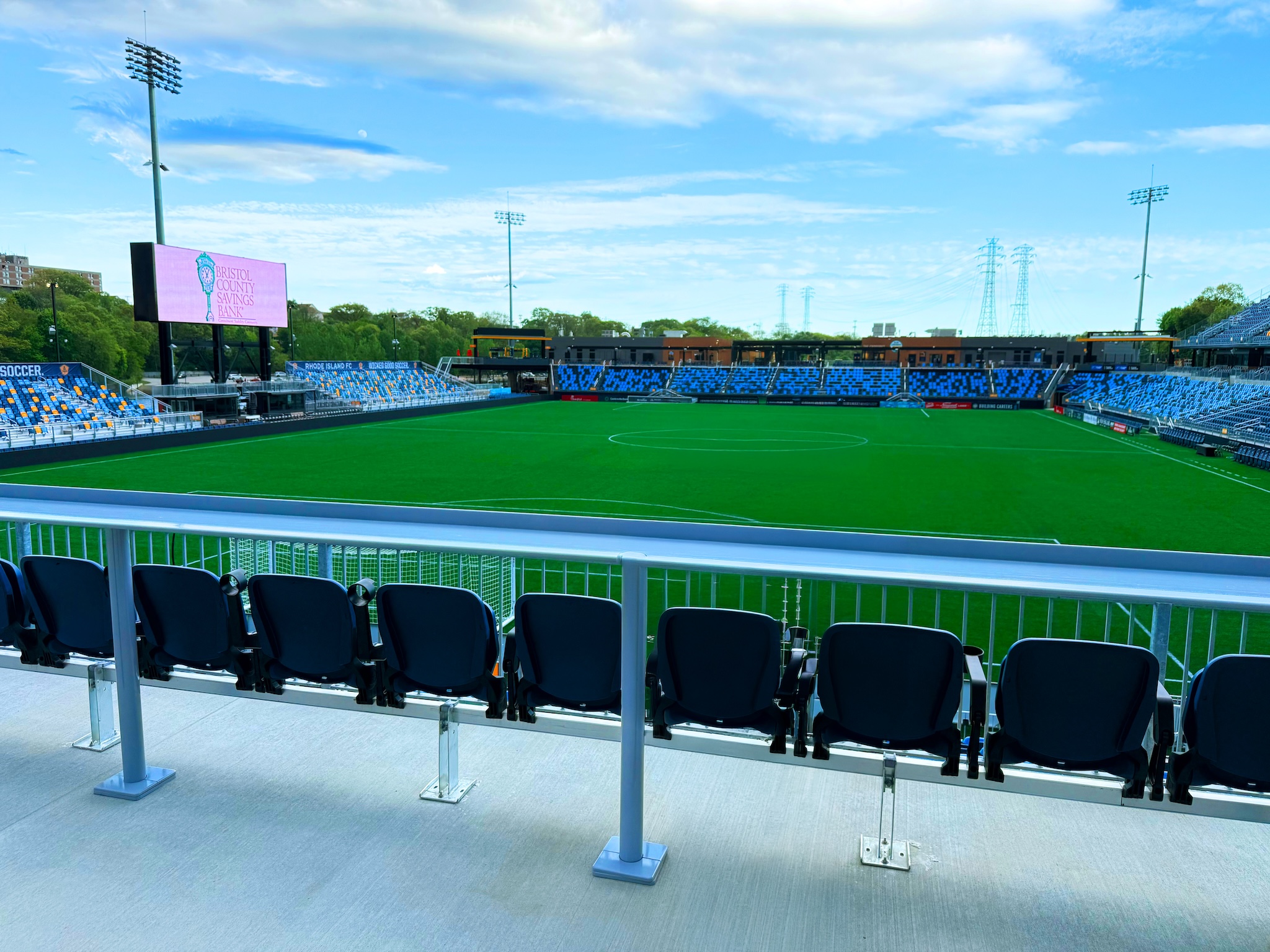 Empty stadium seating with blue and orange chairs facing a green soccer field under a blue sky with clouds, and a large video screen at the left edge.