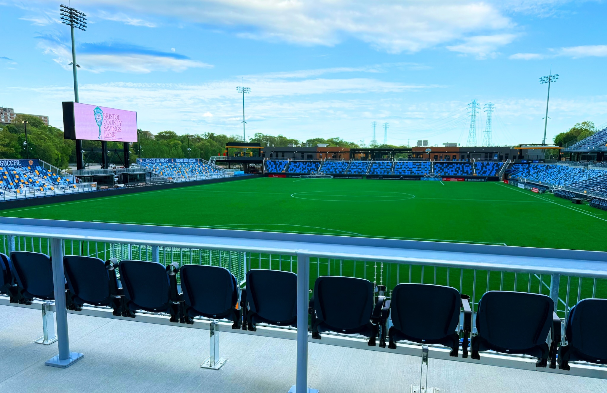 Empty soccer stadium with blue seats, a green field, and a large pink scoreboard on the left edge.