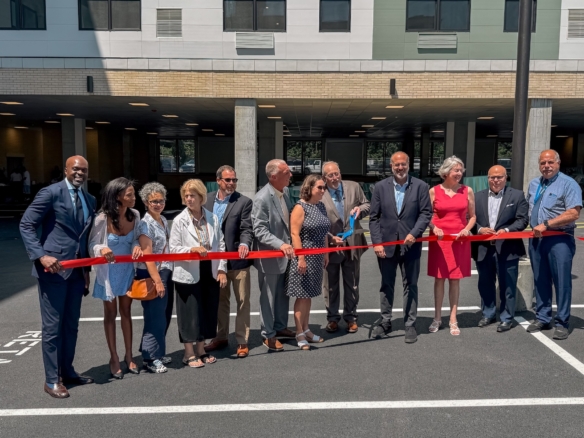 Group of adults in business attire standing behind a red ribbon at a ribbon-cutting ceremony outside a modern building.