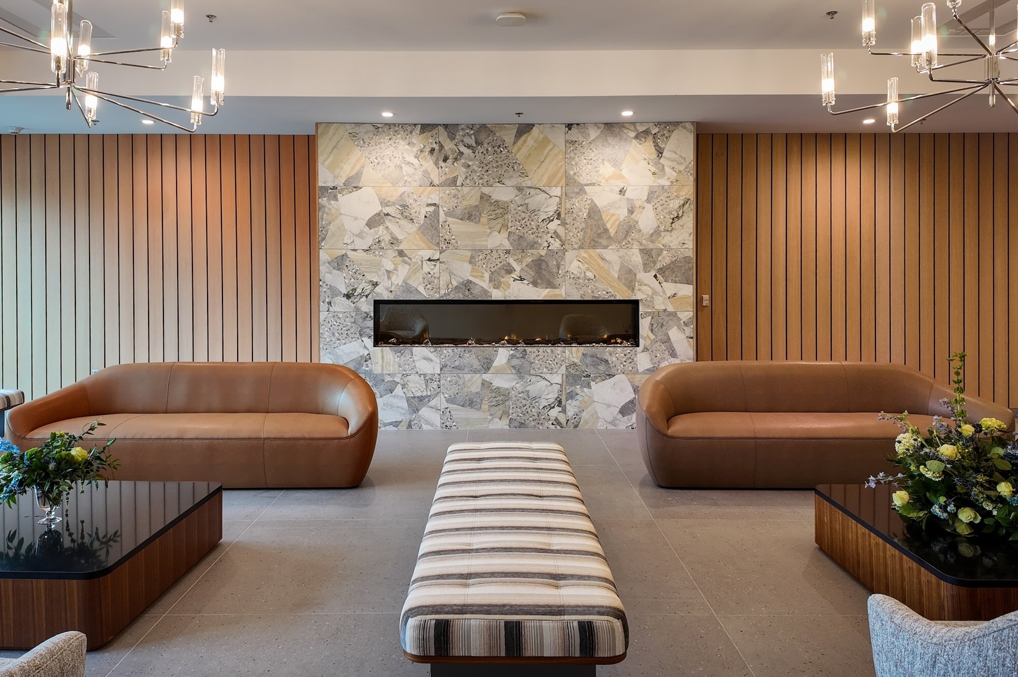 Modern hotel lounge with a mosaic stone fireplace, wood paneled walls, and tan leather couches facing the center bench ottoman.