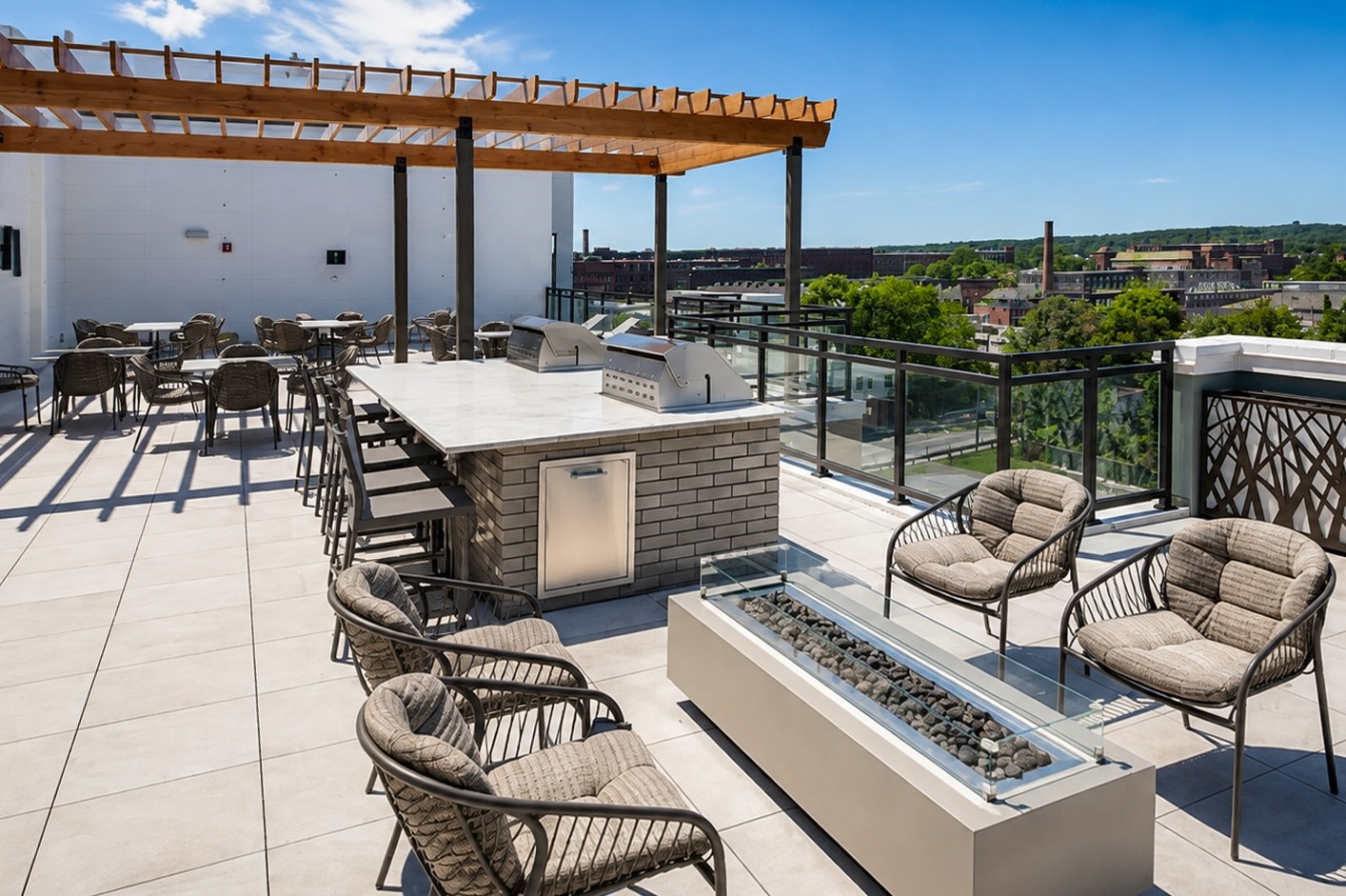 Rooftop terrace with a brick outdoor kitchen and grilling stations, surrounded by lounge chairs and a fire pit, overlooking the city.